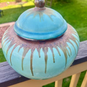 May include: A ceramic jar with a lid, featuring a turquoise glaze with brown drips. The jar has a rounded shape and sits on a wooden railing. The lid is also turquoise with a brown knob.