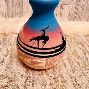 May include: A hand-painted ceramic vase with a blue top and a pink and red body. The vase features a black silhouette design of figures and geometric patterns. The vase is approximately 8 inches tall.