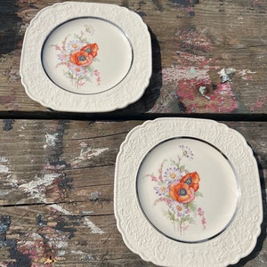 May include: Two white ceramic square plates with a floral design featuring orange poppies and white daisies. The plates have a silver rim and a raised embossed design around the edge.