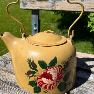 May include: A vintage yellow metal teapot with a wooden handle and a floral design. The teapot features a large red and white rose with green leaves and red buds. The handle and spout are curved.