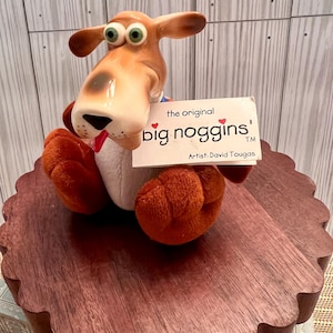 Puede incluir: Un juguete de peluche de perro marrón con ojos grandes y una lengua roja, sosteniendo una tarjeta que dice "the original big noggins! Artist: David Tougas TM".