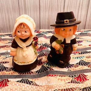 May include: Two ceramic figurines of a girl and a boy dressed in traditional Pilgrim attire. The girl wears a white bonnet and holds red flowers. The boy wears a black hat and holds a green item. Both have smiling faces.