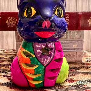 May include: A vibrant, hand-painted ceramic cat sculpture. The cat features a deep blue head with yellow eyes and a red mouth. The body is segmented with bright colors including green, orange, purple, and pink. The sculpture is on a patterned surface.