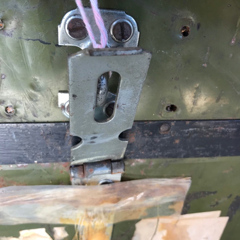 Puede incluir: Primer plano de un pestillo de metal con una bisagra y una placa de metal. El pestillo est&aacute; unido a una superficie de metal verde con varios agujeros. Una cuerda rosa est&aacute; enhebrada a trav&eacute;s del pestillo.