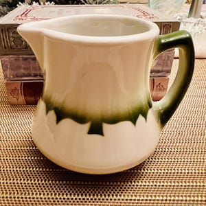 May include: A white ceramic pitcher with a green rim and handle. The pitcher has a green design on the front.