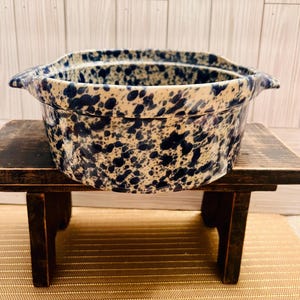 May include: A round, ceramic casserole dish with a speckled blue and cream pattern. The dish has two handles and is sitting on a small, dark wooden table. The table is on a woven, beige mat.