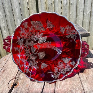 May include: A ruby red glass serving dish with a silver rim and handles. The dish features a floral design and the text "40th Anniversary" in silver. The dish is on a wooden surface.