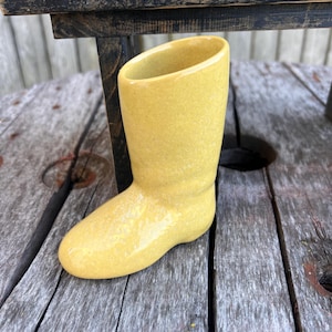 May include: A small, yellow ceramic boot-shaped vase. The vase has a glossy finish and a slanted opening. It is sitting on a weathered wooden surface, with a black wooden structure in the background.