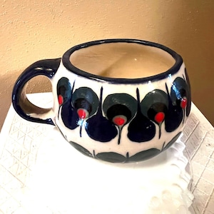 May include: A white ceramic mug with a blue and green floral design. The mug has a rounded shape and a handle.