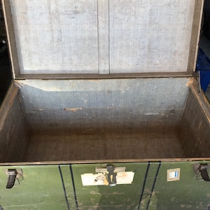 Puede incluir: Un ba&uacute;l de almacenamiento de madera verde vintage con detalles met&aacute;licos negros y una etiqueta en la parte delantera. El ba&uacute;l est&aacute; abierto y el interior est&aacute; forrado con una tela marr&oacute;n claro.