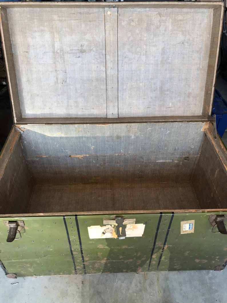 Puede incluir: Un ba&uacute;l de almacenamiento de madera verde vintage con detalles met&aacute;licos negros y una etiqueta en la parte delantera. El ba&uacute;l est&aacute; abierto y el interior est&aacute; forrado con una tela marr&oacute;n claro.