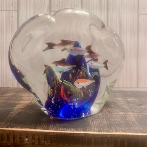 May include: A glass paperweight with a blue base and clear glass top. Inside the paperweight are colorful glass fish and a small amount of green and gold glitter.