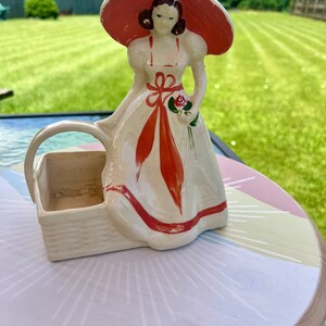 May include: A white ceramic figurine of a woman wearing a red hat and dress, holding a rose. The figurine is holding a white ceramic basket.