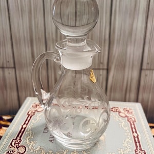 May include: A clear glass decanter with a round stopper and a handle. The decanter features etched floral designs and a gold-colored label. The decanter is sitting on a book with a decorative cover.