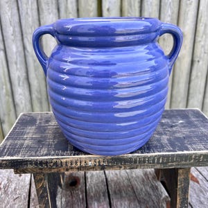 May include: A cobalt blue ceramic vase with two handles. The vase has a ribbed texture and sits on a small, distressed black wooden stool. The background is a weathered wooden fence.