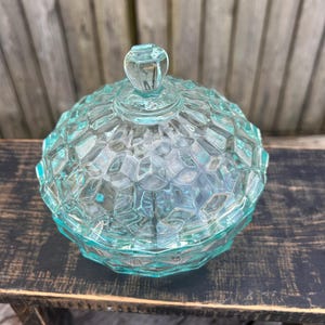 May include: A clear, light blue glass candy dish with a domed lid and a diamond pattern. The dish is sitting on a dark wooden surface.