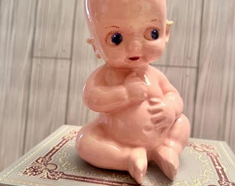May include: A pink ceramic Kewpie doll figurine with blue eyes and a small tuft of hair. The doll is seated, with its hands near its chest. The figurine is on top of a decorative book.