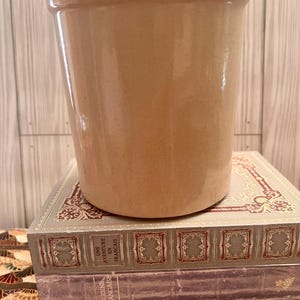 May include: A tan-colored, cylindrical stoneware crock. The crock is sitting on top of a stack of vintage books with decorative covers. The books have French titles, suggesting a culinary or literary theme.