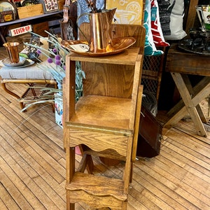May include: A wooden step stool with three steps, the top step is a shelf. The stool is made of light brown wood with a wavy edge on the top step. A copper pitcher and a copper tray are on the top shelf. The stool is in a room with a wooden floor.