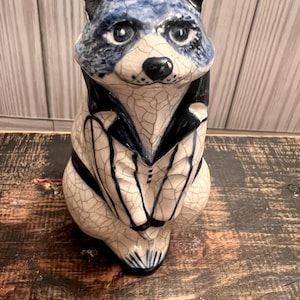 May include: A ceramic raccoon figurine with a blue and white color scheme. The raccoon is depicted wearing a dark blue tuxedo. The figurine has a crackle glaze finish and is posed in a seated position.