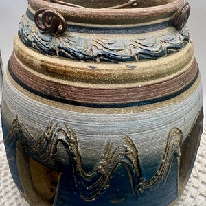 May include: A brown, white, and blue ceramic pot with a wavy design. The pot has two metal loops on the top.