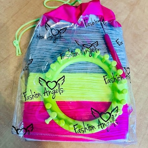 May include: A set of colorful yarn in a clear plastic bag with a pink drawstring bag. The yarn is in shades of pink, yellow, and gray. The bag also includes a green plastic loom and a crochet hook. The bag has the words "Fashion Angels" printed on it.