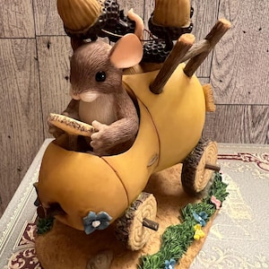 May include: A figurine of a brown mouse driving a yellow acorn car with two other mice riding in the back. The car is on a base with green grass and blue flowers.