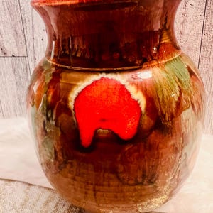 May include: A handcrafted ceramic vase with a glossy finish. The vase features a rich brown base with streaks of green and a prominent red accent. The vase has a wide body and a flared rim, showcasing a unique, artistic design.