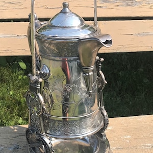 May include: A silver plated tea or coffee pot with an ornate design and a handle. The pot has a base with a small bowl attached to it.