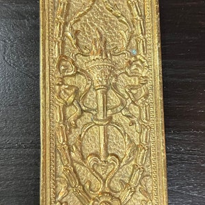 May include: A gold-toned metal door plate with an ornate design featuring a central motif of a torch with flames. The plate has a rectangular shape with rounded corners and a decorative border.
