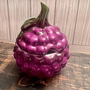 May include: A purple ceramic grape-shaped serving dish with a lid. The dish is made to look like a bunch of grapes.