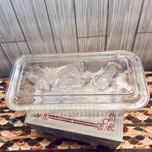 May include: A clear, rectangular glass casserole dish with a lid, featuring embossed corn designs. The dish is resting on a decorative box with a patterned design. The background is a light wood panel.