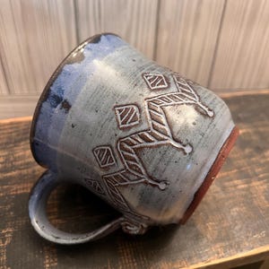 May include: A blue ceramic mug with a brown geometric design. The mug has a handle and is sitting on a wooden surface.