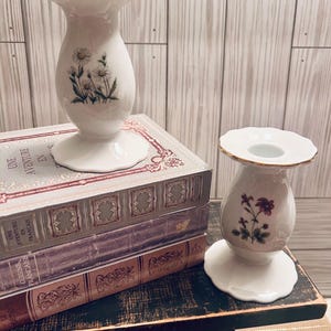 May include: Two white ceramic candle holders with floral designs. The holder on the left has white flowers and the holder on the right has red flowers. The holders are sitting on top of a stack of books.
