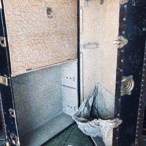 May include: An antique black and brown vintage suitcase with a floral patterned interior. The suitcase is open and has a metal hanger inside. The suitcase is sitting on a green surface.