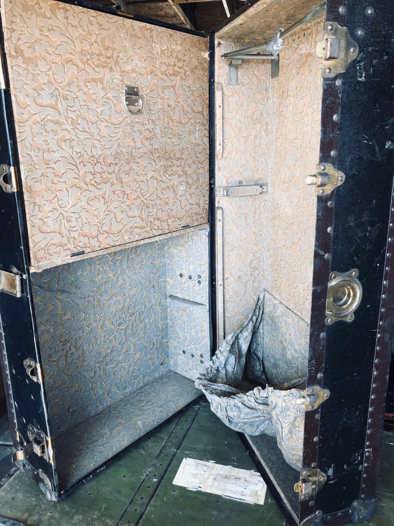 May include: An antique black and brown vintage suitcase with a floral patterned interior. The suitcase is open and has a metal hanger inside. The suitcase is sitting on a green surface.