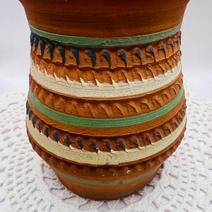 May include: A brown terracotta pot with a decorative design of green, white, and black stripes. The pot has a rough, textured surface and a slightly flared rim.
