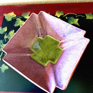 May include: A square ceramic dish with a deep green center and a purple and pink glaze. The dish has a geometric design and is perfect for serving snacks or small items.