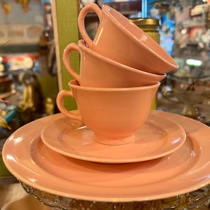 May include: A stack of four light pink teacups and saucers. The cups have curved handles and sit atop two plates. The set is displayed on a decorative glass stand, with a blurred background of a shop interior.