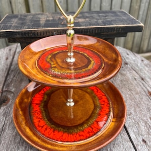 May include: A two-tiered serving tray with a gold-colored handle. The ceramic plates are brown with a vibrant red and orange circular design. The tray is likely vintage and suitable for serving desserts or appetizers.