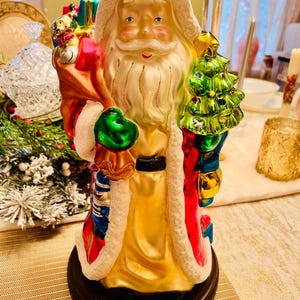 May include: A glass figurine of Santa Claus holding a green Christmas tree and a bag of gifts. Santa is wearing a red and white suit with a gold trim and a white beard. The figurine is on a black base.