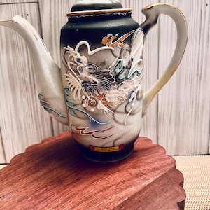 May include: A black and white ceramic teapot with a gold trim and a dragon design. The teapot has a white spout and handle. The teapot is sitting on a wooden base.