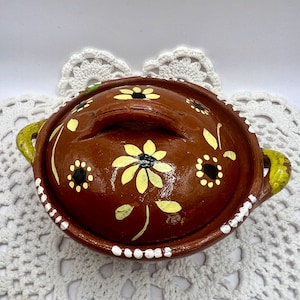 May include: A small brown clay pot with a lid, decorated with yellow and black flowers. The pot has two yellow handles and a white rim.