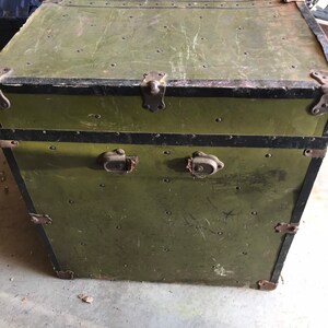 Puede incluir: Un ba&uacute;l vintage de estilo militar verde con ribetes negros y cierres met&aacute;licos. El ba&uacute;l tiene un aspecto desgastado y deteriorado con ara&ntilde;azos y abolladuras visibles.