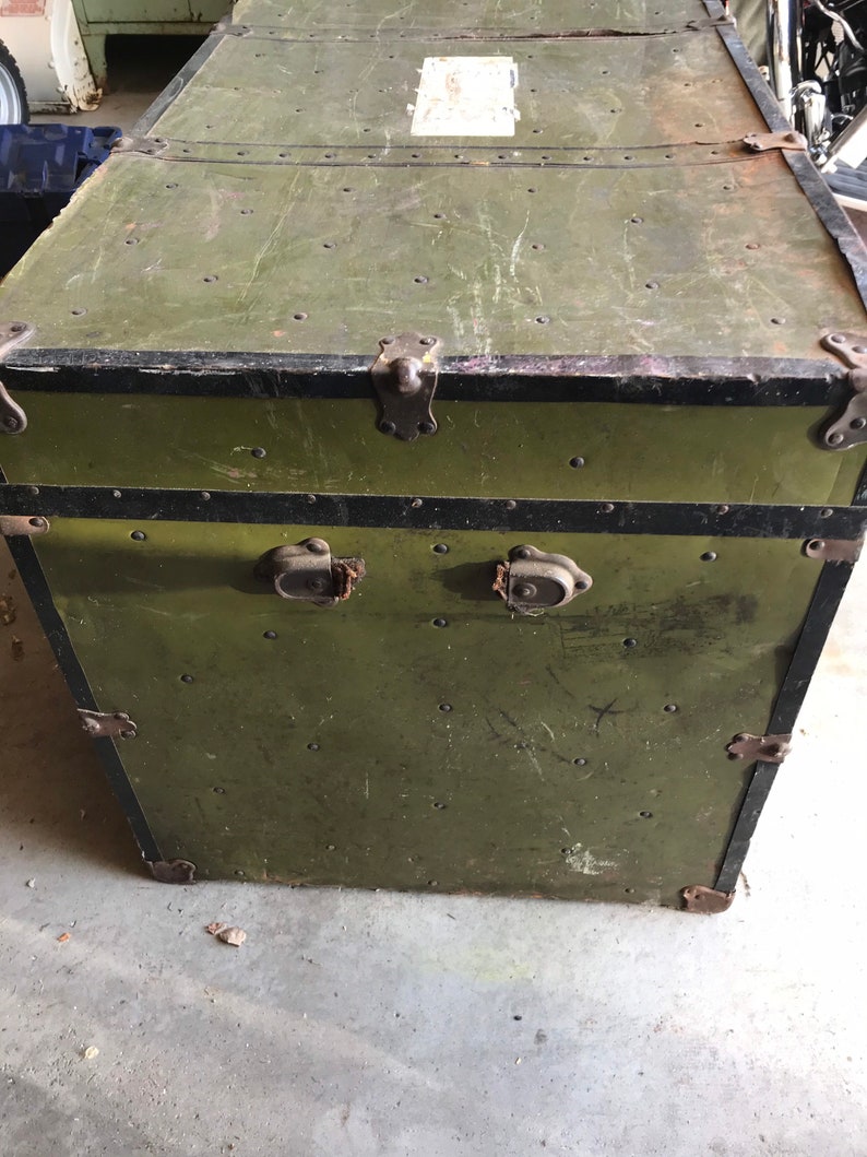 Puede incluir: Un ba&uacute;l vintage de estilo militar verde con ribetes negros y cierres met&aacute;licos. El ba&uacute;l tiene un aspecto desgastado y deteriorado con ara&ntilde;azos y abolladuras visibles.