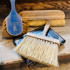 May include: An assortment of vintage paint brushes and a scrub brush. The brushes have wooden handles and natural bristles. One brush is blue with the text "504 NORCRAFT". Another brush is labeled "12" BESTRUY". The brushes are resting on a wooden surface.