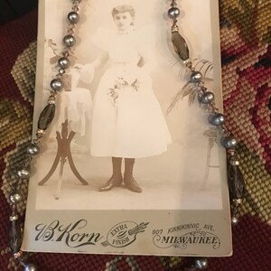 May include: A vintage photograph of a young woman wearing a white dress and holding a bouquet of flowers. The photo is framed with a beaded necklace with gray and brown beads. The photo is signed "W.B. Korn, Extra Finish, 907 Kinnickinnic Ave., Milwaukee."