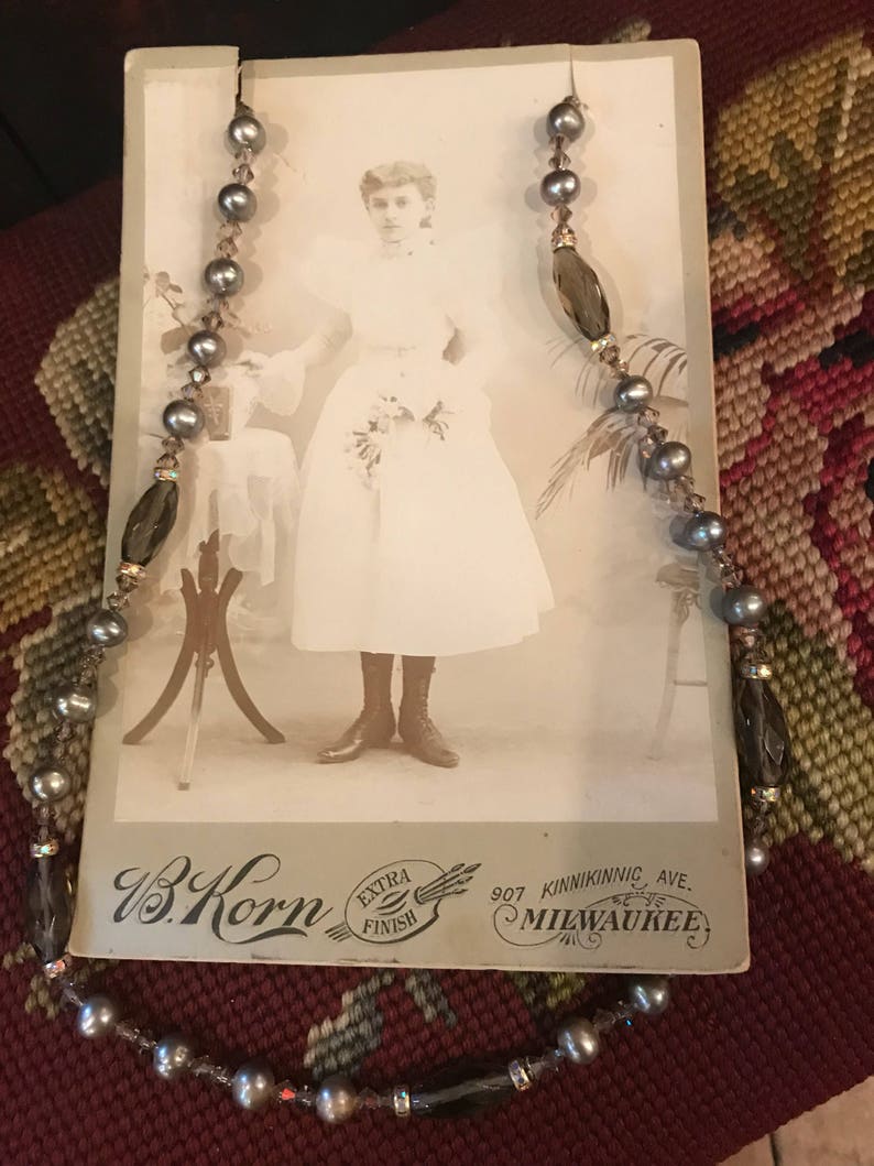 May include: A vintage photograph of a young woman wearing a white dress and holding a bouquet of flowers. The photo is framed with a beaded necklace with gray and brown beads. The photo is signed "W.B. Korn, Extra Finish, 907 Kinnickinnic Ave., Milwaukee."
