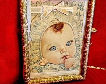 May include: A vintage picture frame with a gold and pink finish. The frame contains a picture of a baby with a white bonnet and a bow. The baby is looking at the viewer and has a finger in its mouth.
