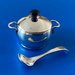 May include: Silver serving set with a small covered pot and a ladle. The pot has a black knob on the lid.